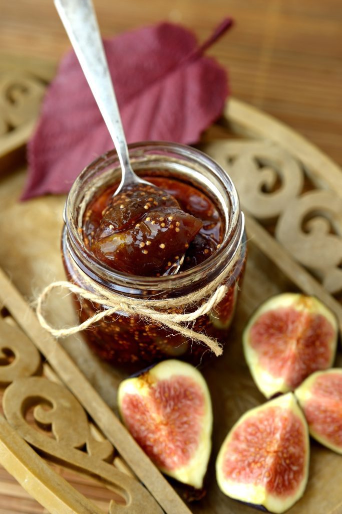 Jar of fig jam with a spoon and fresh fig halves on a wooden tray.