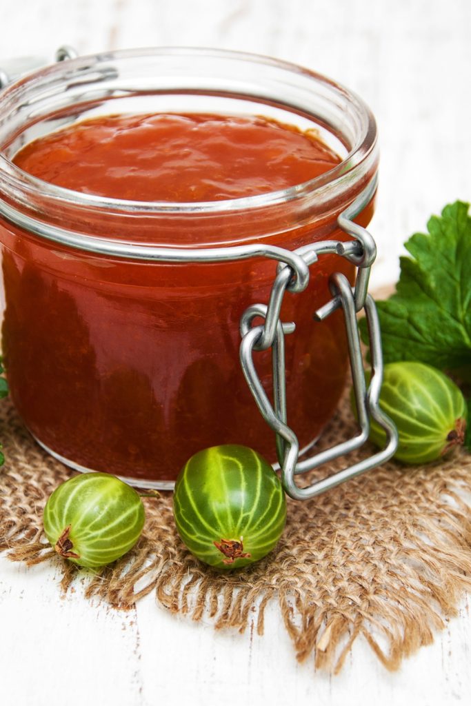 Jar of gooseberry jam with fresh green gooseberries on a rustic cloth.