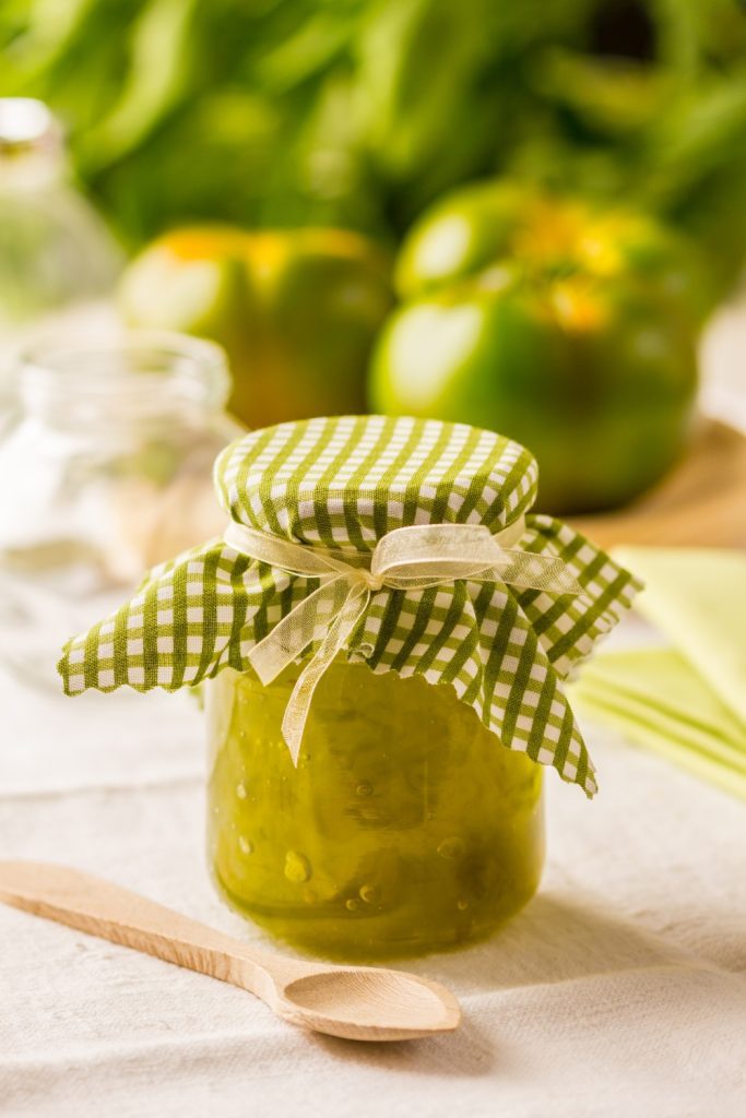 Jar of Slow Cooker Green Tomato Jam with green tomato halves and a spoon on a crispbread.