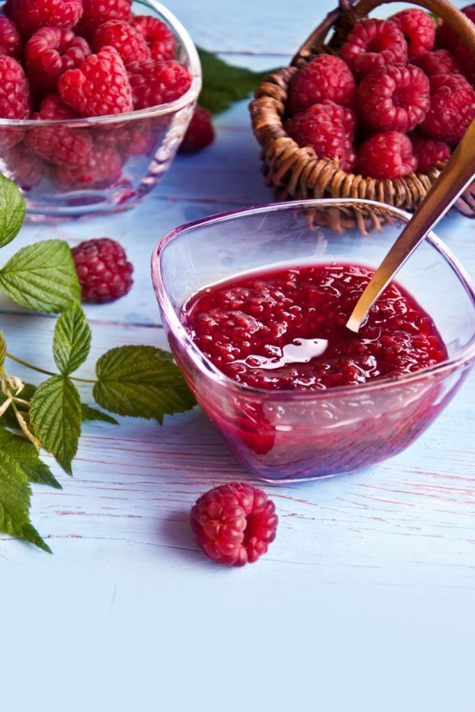 Jar of raspberry jam with fresh raspberries on a wooden surface.