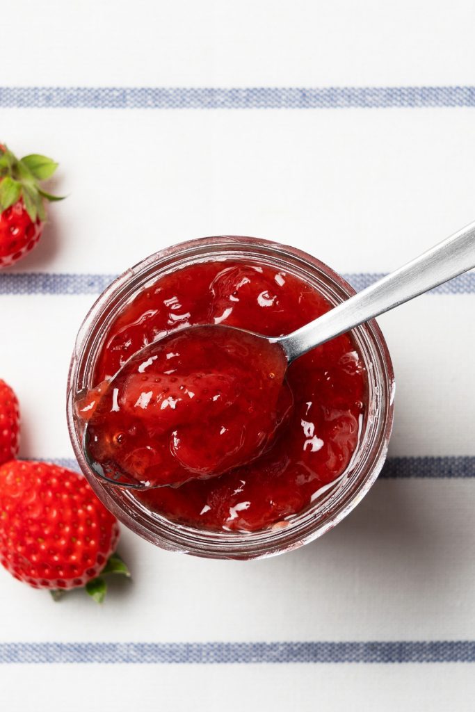 Jar of strawberry jam with a spoon on a striped cloth and fresh strawberries beside.