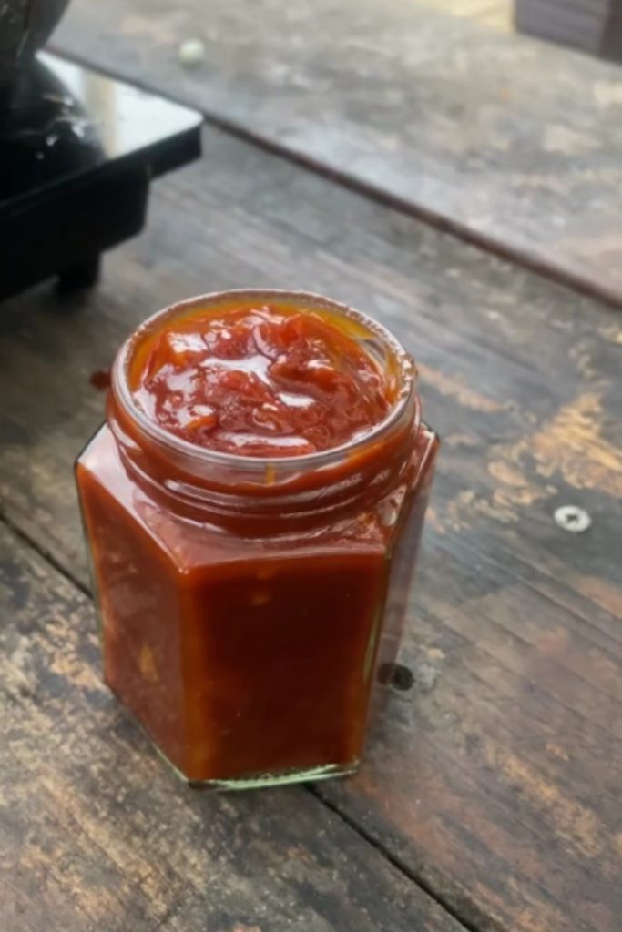 Tomato Capsicum Relish in a hexagonal glass jar on a wooden surface.
