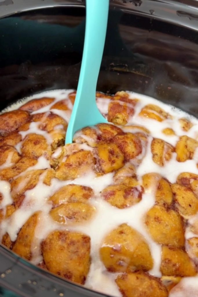 apple cinnamon rolls in a crockpot drizzled with icing, with a blue spatula inserted.