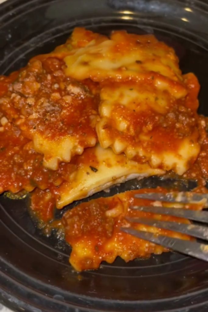 A fork cutting into crockpot ravioli with tomato sauce in a black bowl.