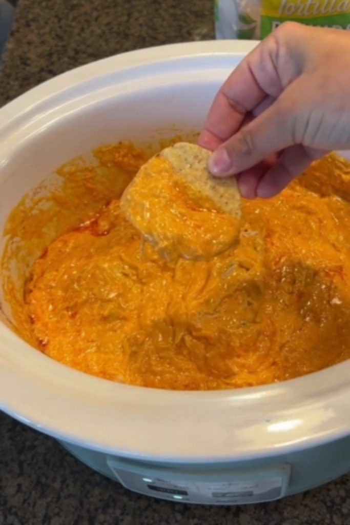 A hand dipping a tortilla chip into 5 Ingredient Crock Pot Buffalo Chicken Dip in a slow cooker.