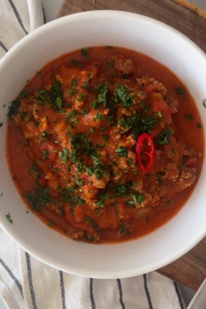 A white bowl filled with 5 Ingredient No Bean Crockpot Chili, topped with shredded cheese and sliced red chili.