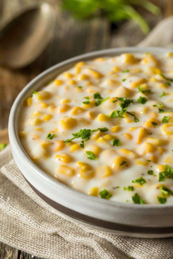 A bowl of creamed corn garnished with parsley on a beige napkin.