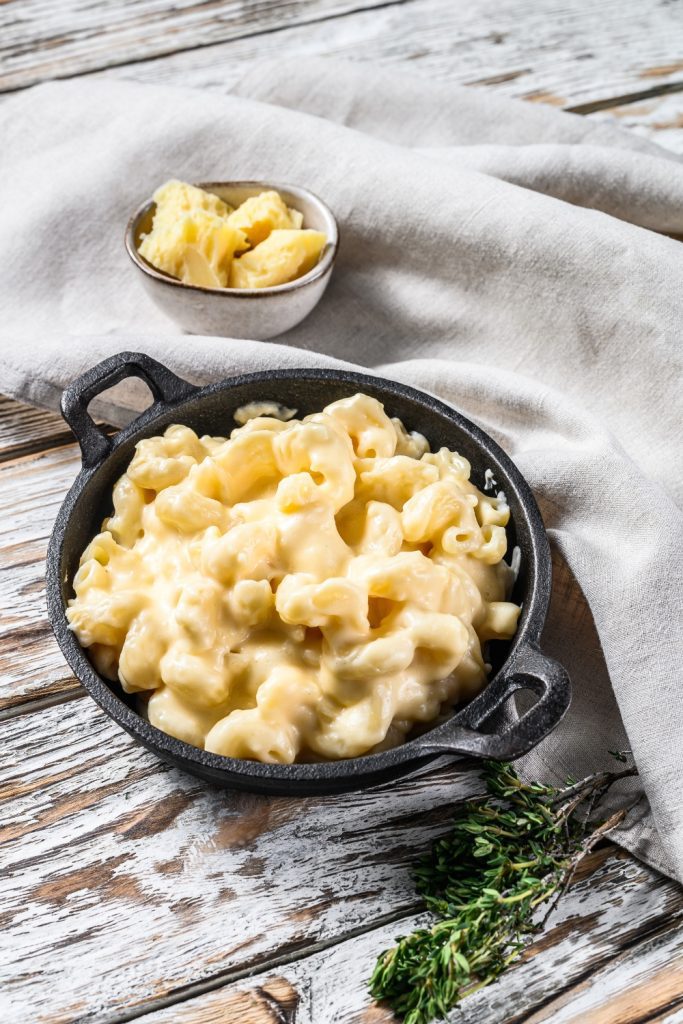 A cast iron skillet filled with creamy 5 Ingredient Slow Cooker Mac and Cheese on a wooden table, with a small bowl of cheese pieces on the side.