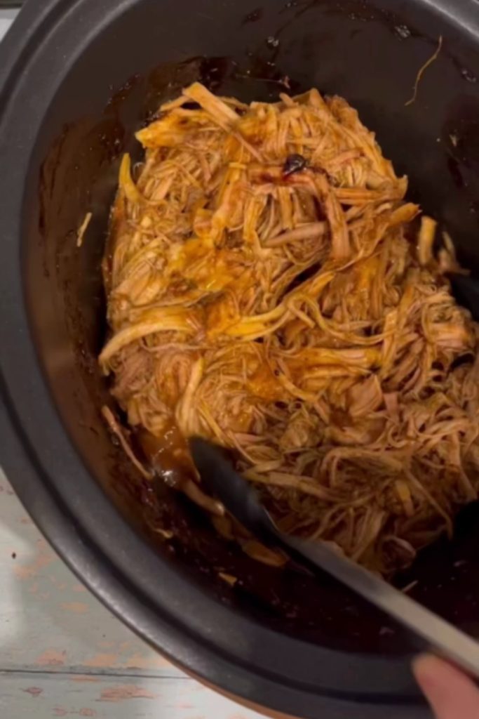 Shredded pulled pork in a slow cooker with tongs.