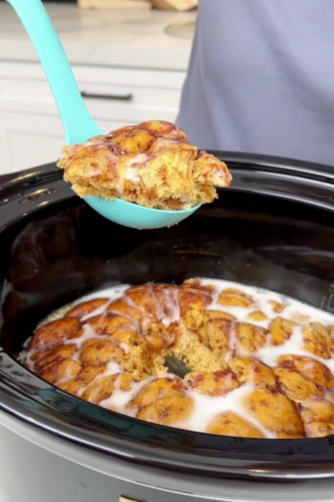 A serving of apple cinnamon rolls being lifted out of the crockpot with a blue spatula.