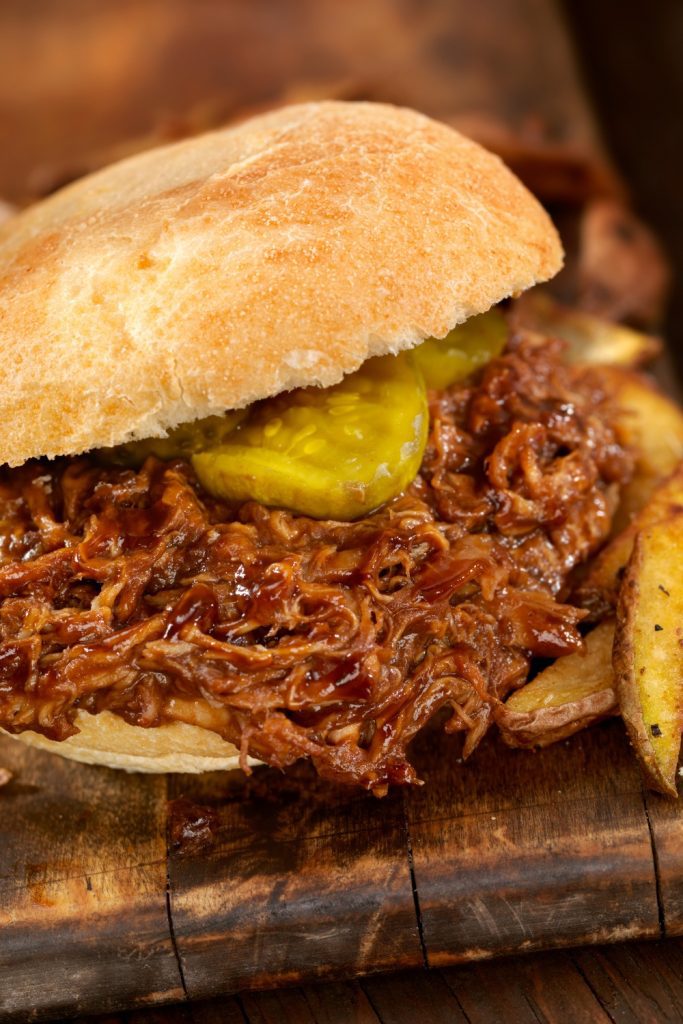 Pulled pork sandwich with pickles on a bun, served with potato wedges.