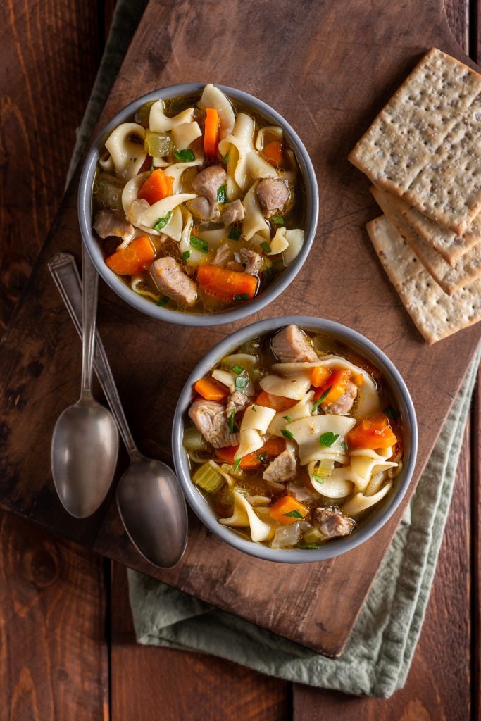 Two bowls of chicken noodle soup with crackers on the side.