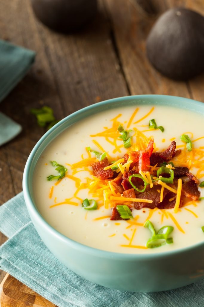 Potato soup with bacon, cheese, and green onions in a blue bowl on a wooden table.