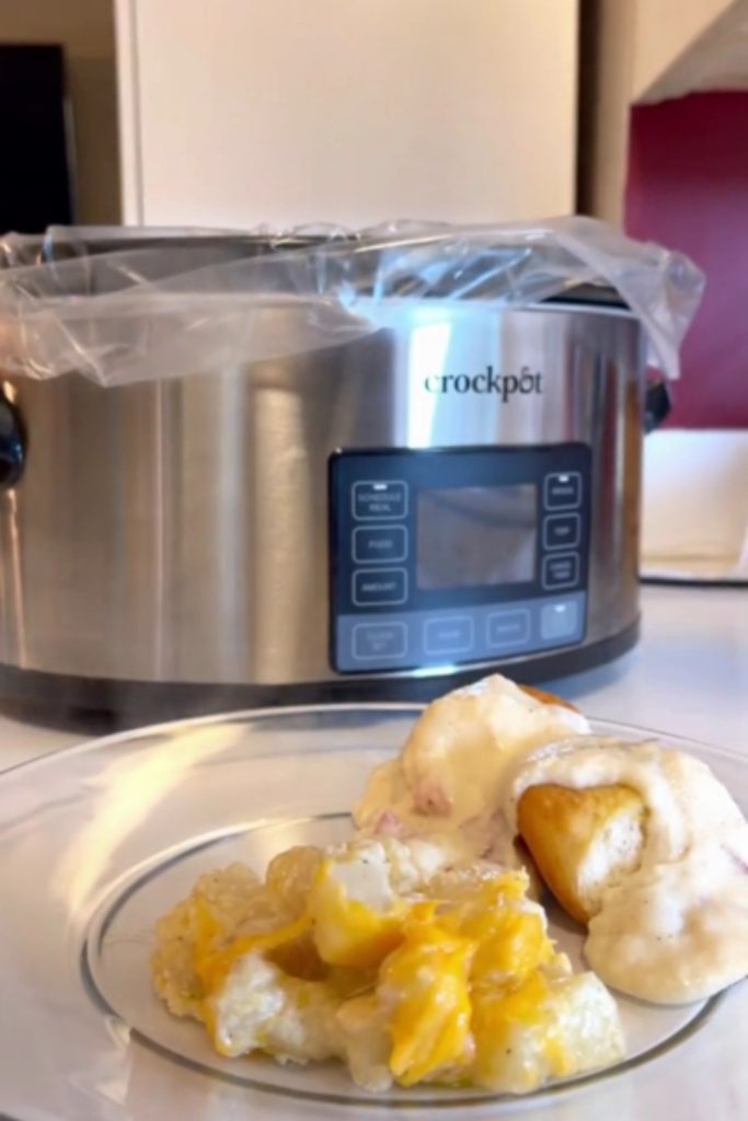 Plate of cheesy potatoes and a bread roll in front of a slow cooker.