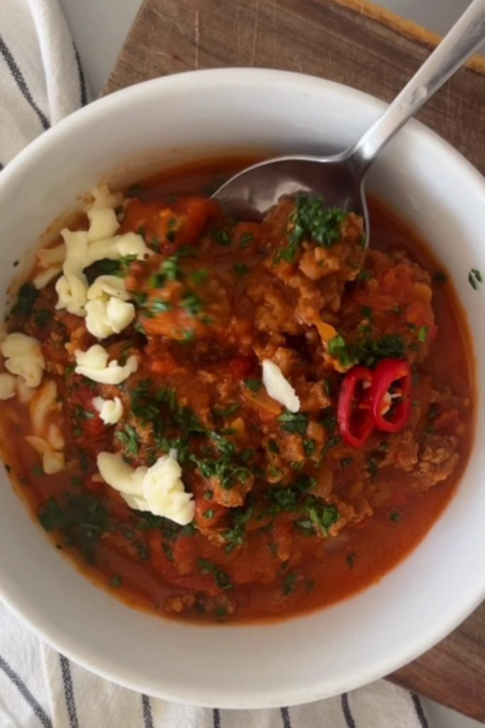 A spoonful of 5 Ingredient No Bean Crockpot Chili held over a white bowl filled with the chili, topped with shredded cheese and sliced red chili.