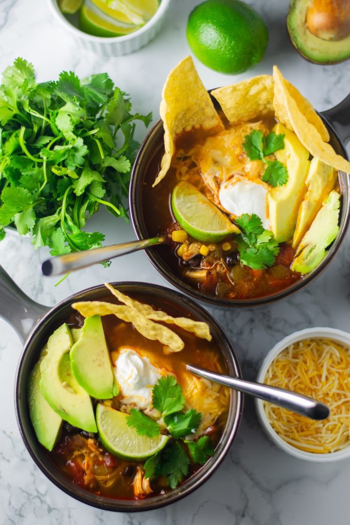 Chicken tortilla soup in two bowls topped with avocado, lime, cilantro, and tortilla chips.